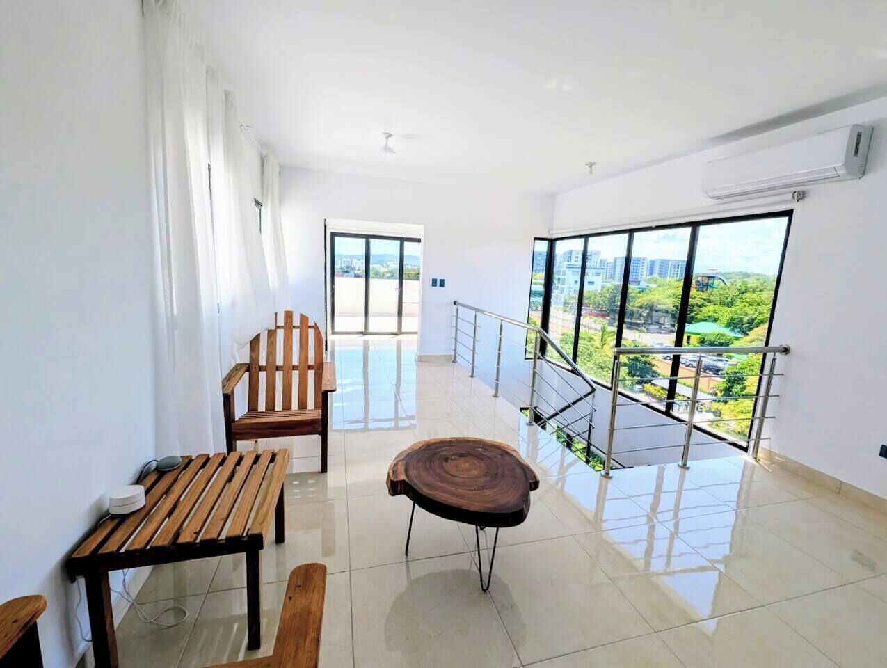 A spacious balcony with a view of greenery and buildings, a wooden bench, a table, and a unique wooden stool.