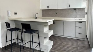 Et kompakt køkken med hvide skabe, en lille ø med barstole og et flisebeklædt bagpanel.