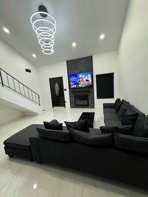 Ein modernes Wohnzimmer mit einem Ecksofa, einem Flachbildfernseher an der Wand, einem Kamin und einer Treppe, die in eine obere Ebene führt.