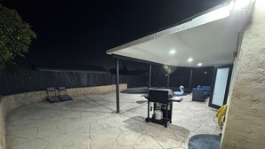 En overdækket patio med grill, udendørs møbler og swimmingpool.