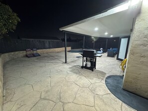 Ein überdachter Patio-Bereich mit Grill, Gartenmöbeln und einem Swimmingpool.