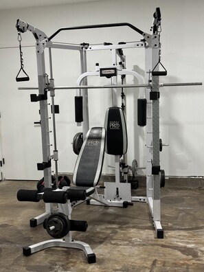 Ein Fitnessraum mit einer multifunktionalen Trainingsstation, darunter eine Bank, ein Squat-Gestell und verschiedene Gewichte.