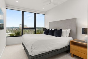 A modern bedroom with a large window, a bed with white bedding, and a grey headboard. - Altura Bay Luxury Residence Frankston (Frankston)