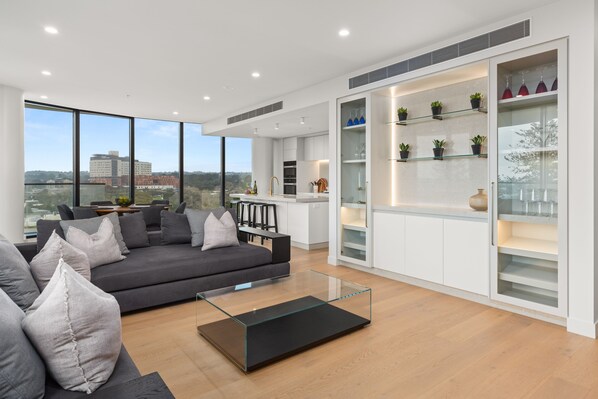A modern living room with a grey sofa, a glass coffee table, and a view of the city through large windows.