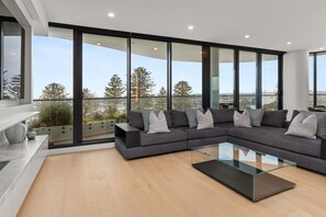 Una sala de estar moderna con un sofá seccional, una mesa de centro de vidrio y ventanas amplias que ofrecen vistas a árboles y edificios.