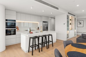 A modern kitchen with white cabinetry, stainless steel appliances, and a central island with bar stools. - Altura Bay Luxury Residence Frankston (Frankston)