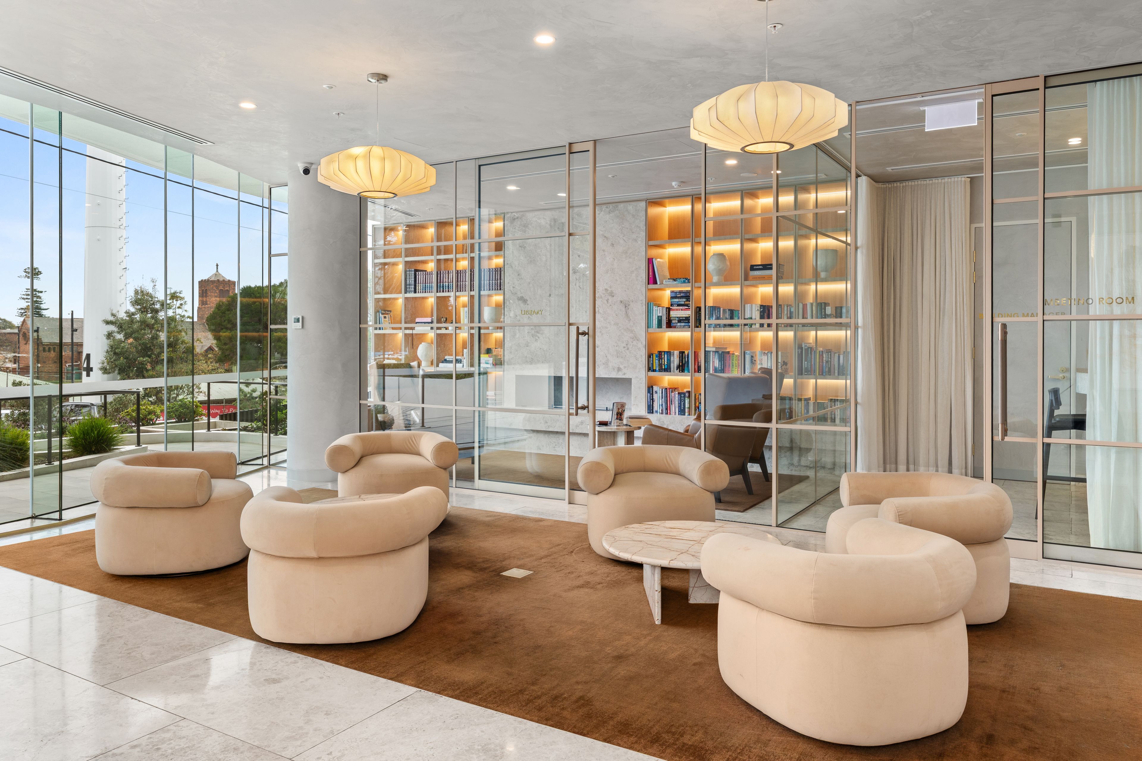 A modern lobby with a glass wall, beige sofas, a marble floor, and a bookshelf with books and decorative items.