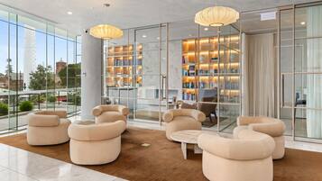 A modern lobby with a glass wall, beige sofas, a marble floor, and a bookshelf with books and decorative items.