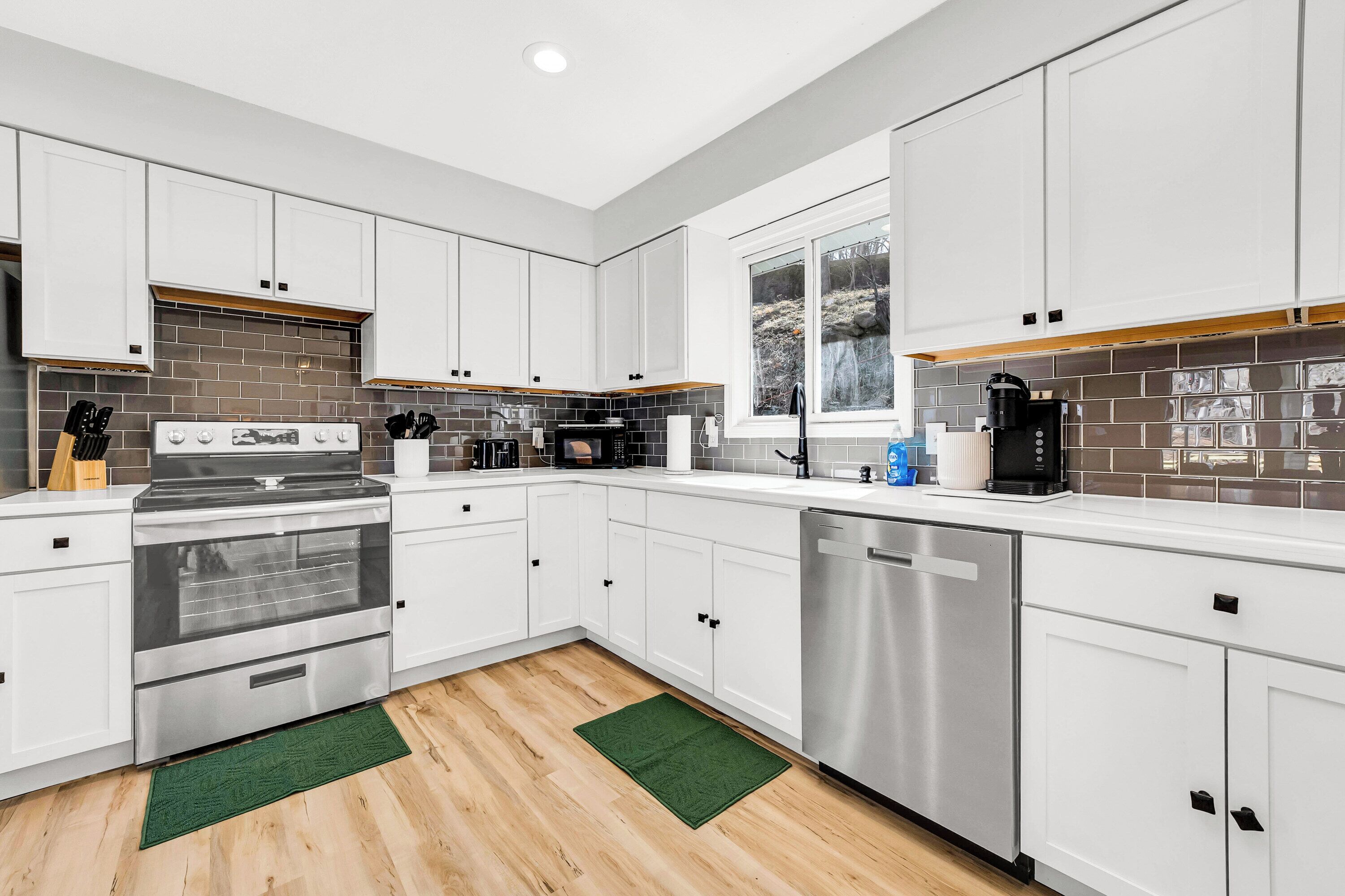 A modern kitchen with white cabinets, stainless steel appliances, and a window with a view of trees.