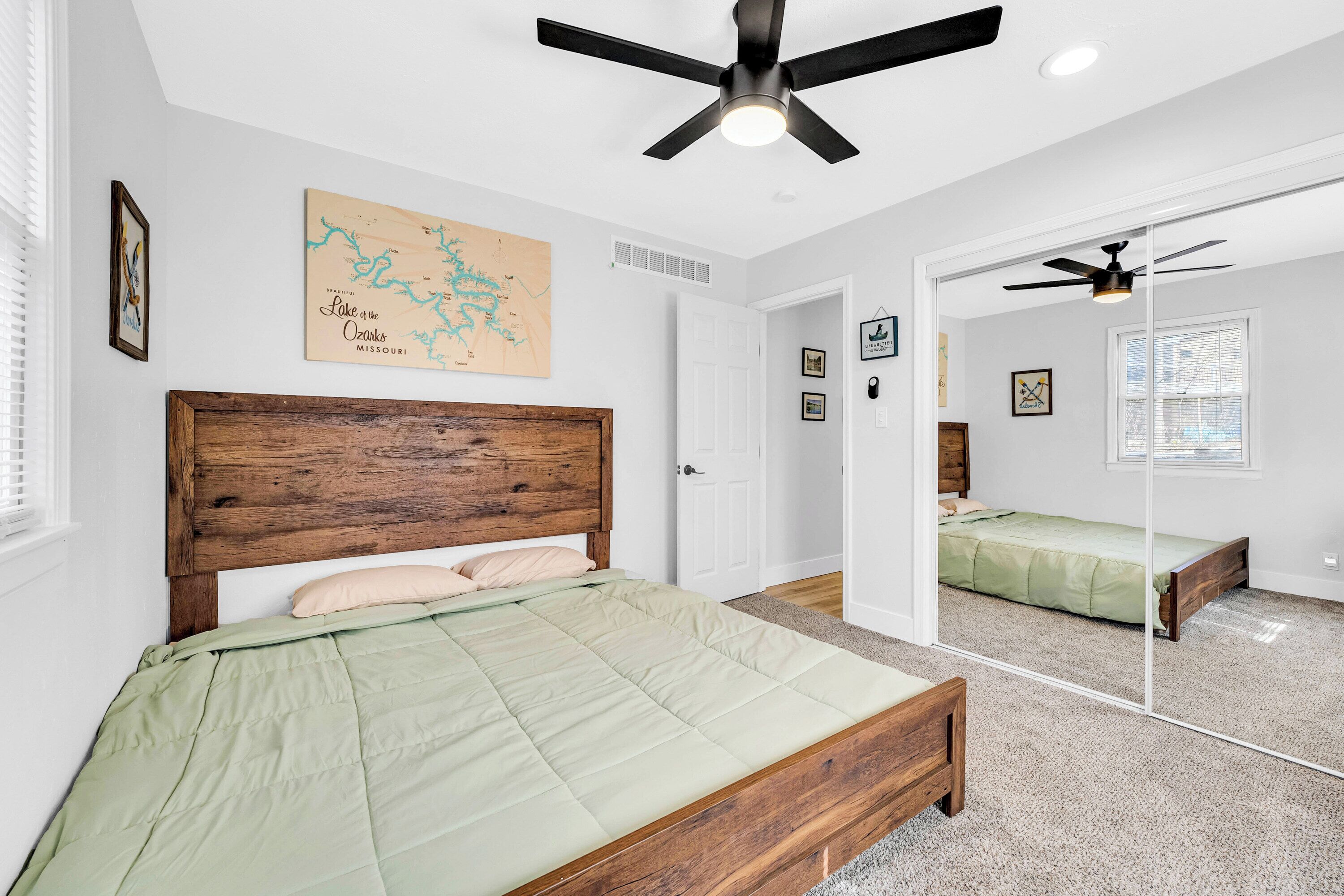A bedroom with a wooden headboard, a green bedspread, a ceiling fan, a map on the wall, and a large mirror reflecting the room.