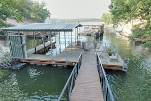 Une jetée en bois avec un espace de repos abrité et un petit bâtiment, probablement des toilettes, sur une rivière paisible, avec des arbres et un pont en arrière-plan.