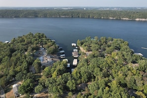 Une vue aérienne d’une propriété au bord du lac, où plusieurs bateaux sont amarrés, le tout entouré d’une forêt dense et de quelques bâtiments.