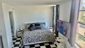 A hotel room with a checkered floor, a bed with patterned bedding, a nightstand with a lamp, a wall-mounted TV, and a window with curtains. - Spacious Studio Apartment Best Location (Miami Beach)