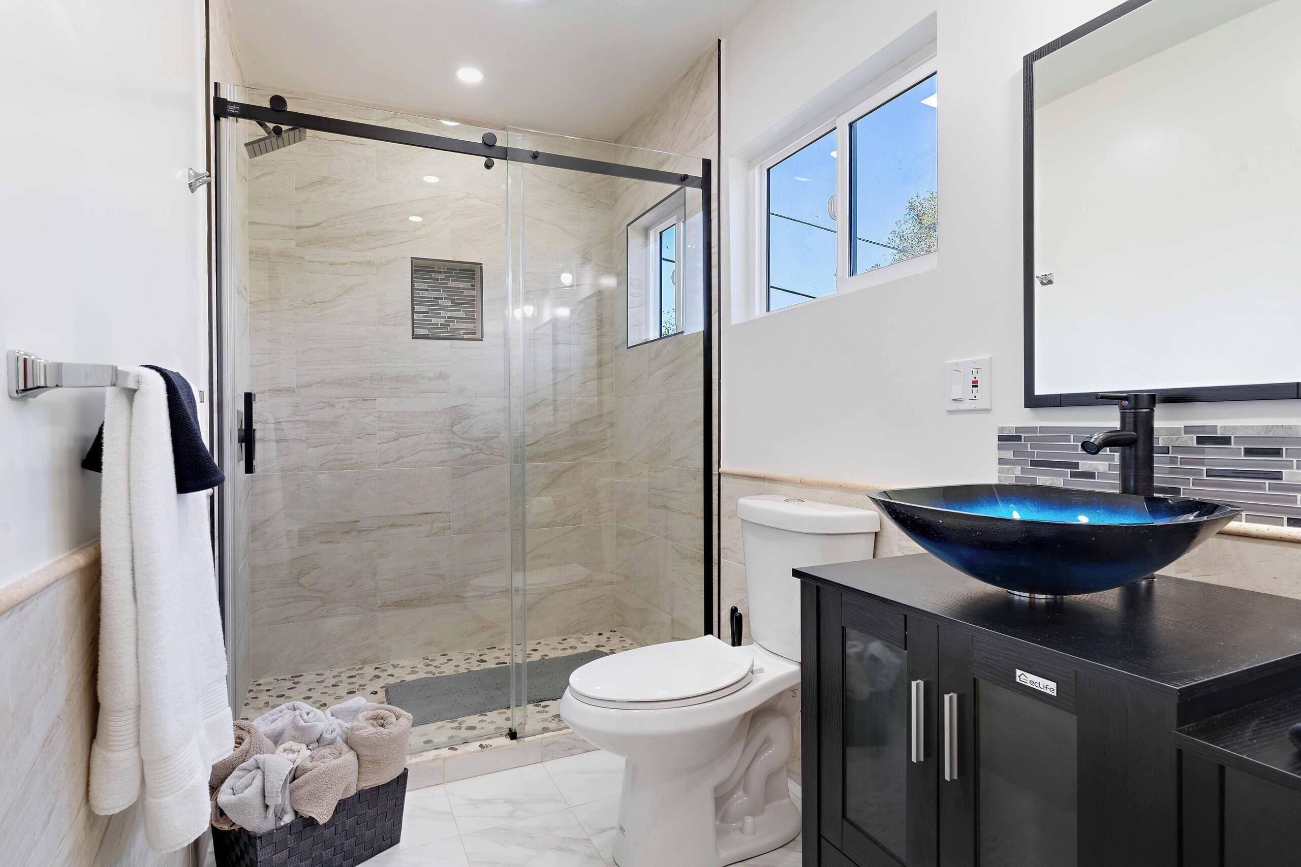Une salle de bain moderne avec une douche à l’italienne en verre, des toilettes blanches et un lavabo noir avec un receveur bleu.
