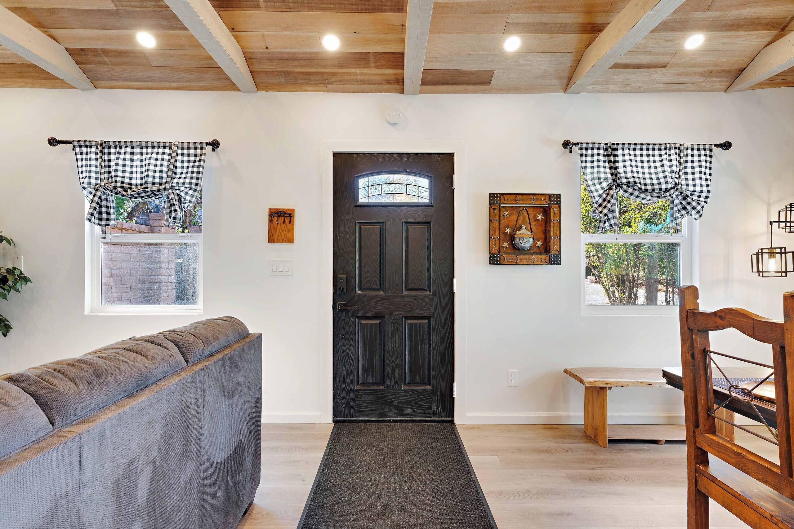 A room with a dark wooden door, checkered curtains, a grey sofa, a small wooden table, and a wooden chair.