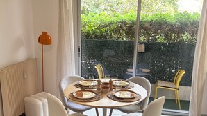 Comedor con una mesa redonda dispuesta para cuatro personas, un radiador, una lámpara de pie y vistas a un jardín verde a través de un ventanal grande.