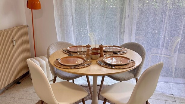 Un comedor con una mesa redonda dispuesta para cuatro personas, rodeada de sillas blancas, un tapete tejido y un radiador.