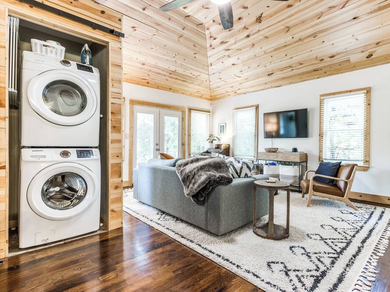A compact living space with a washing machine, a grey sofa, a small table, a TV, and a wooden ceiling.