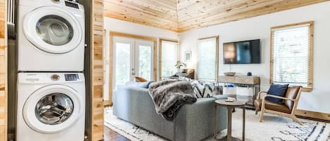 A compact living space with a washing machine, a grey sofa, a small table, a TV, and a wooden ceiling.