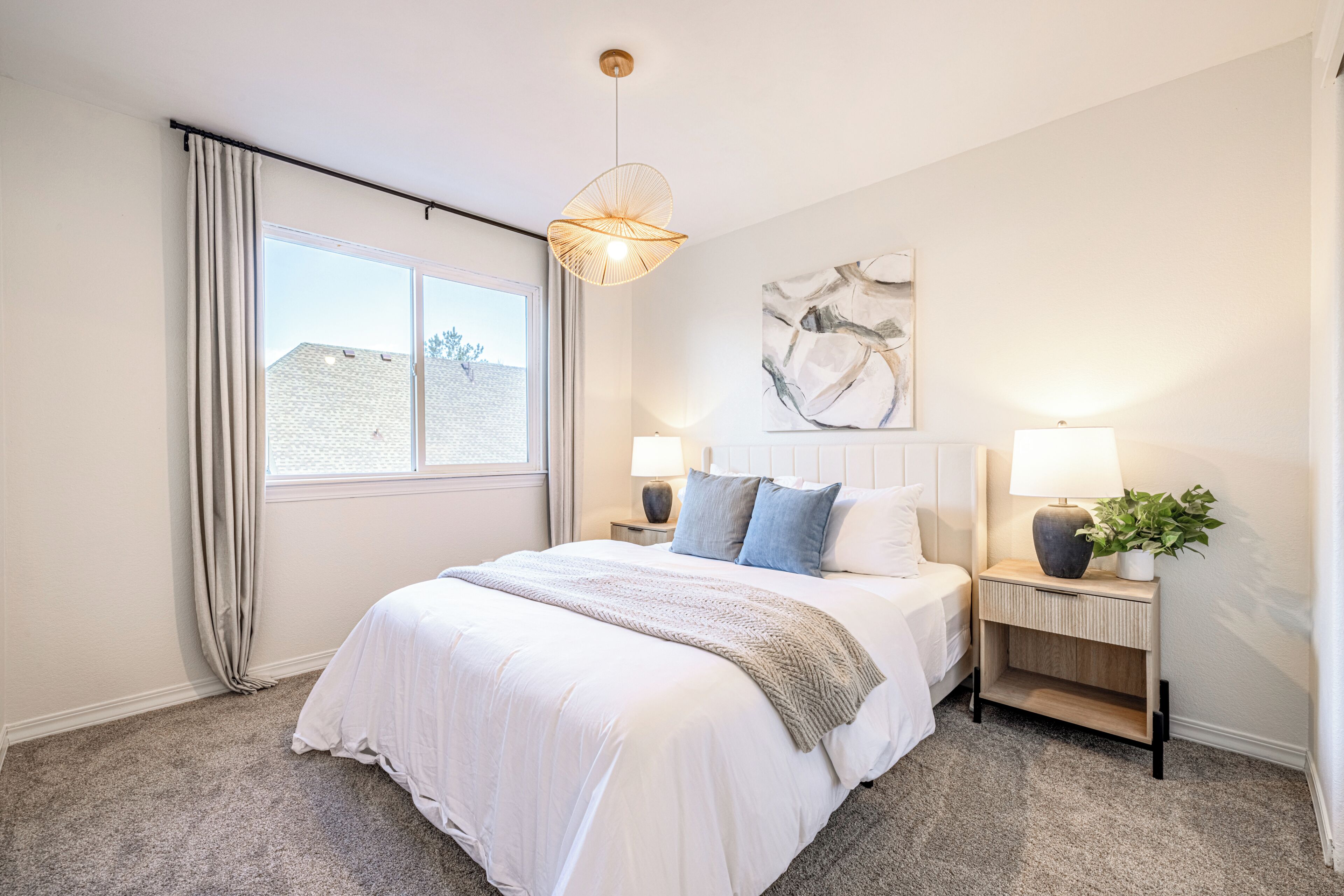 A bedroom with a large bed, two bedside tables with lamps, a window with curtains, and a wall-mounted abstract painting.