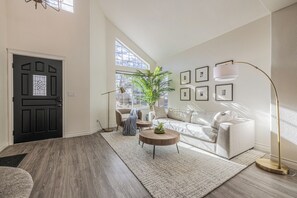 Ein modernes Wohnzimmer mit einer schwarzen Tür, einem großen Fenster, einem weißen Sofa, einem runden Couchtisch und einer Stehlampe.
