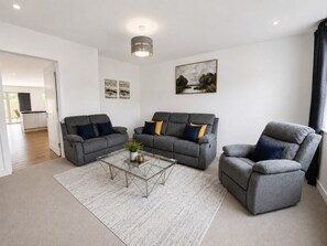 A modern living room with a sofa, armchair, and coffee table, a painting on the wall, and a door leading to another room. - The Riverside • 4 Bedroom 4 Bathroom • New Build House • Sleeps 7 • Driveway Parking • Yoko Property (Huntingdon)