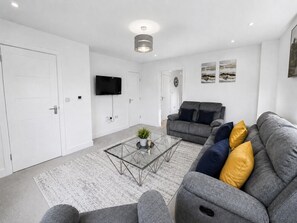 A modern living room with a glass coffee table, grey sofa, and a flat-screen TV mounted on the wall. - The Riverside • 4 Bedroom 4 Bathroom • New Build House • Sleeps 7 • Driveway Parking • Yoko Property (Huntingdon)