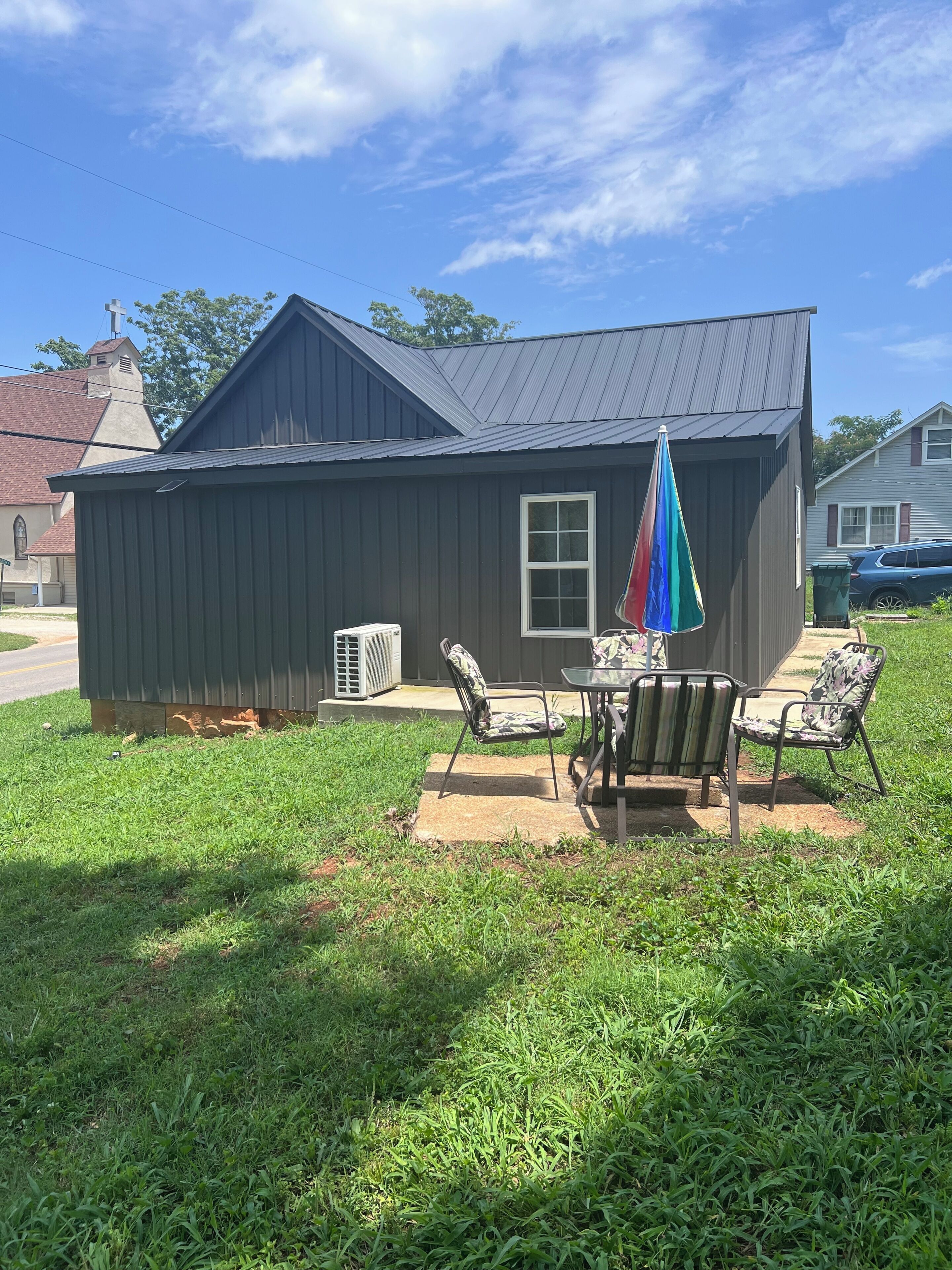 A small building with a metal roof and a window, surrounded by a grassy yard with two chairs and a colorful umbrella.