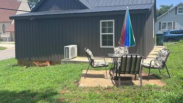 A small building with a metal roof and a window, surrounded by a grassy yard with two chairs and a colorful umbrella.