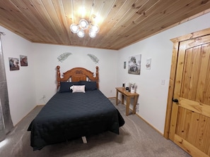 A bedroom with a wooden ceiling, a bed with a dark bedspread, a bedside table with a lamp, and a door. - Cozy 2-bedroom house in Eminence with AC. Located in the heart of town!! (Eminence)
