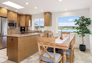 A modern kitchen with a central island, stainless steel appliances, wooden cabinetry, and a view of a lake through large windows. - Whidbey Island Updated Beachfront Getaway - A View From Every Room! (Oak Harbor)