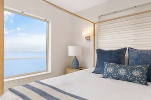 A hotel room with a large window, a bed with blue pillows, a bedside table with a lamp, and a view of the sea. - Whidbey Island Updated Beachfront Getaway - A View From Every Room! (Oak Harbor)