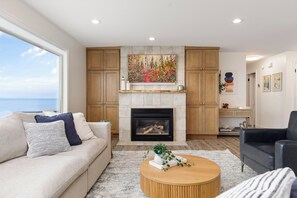 A living room with a fireplace, wooden cabinets, a sofa, a coffee table, and a view of the sea. - Whidbey Island Updated Beachfront Getaway - A View From Every Room! (Oak Harbor)