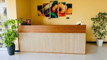Un mostrador de recepción con un panel de rayas verticales, una planta en maceta a la izquierda y una obra de arte floral colgada en la pared.