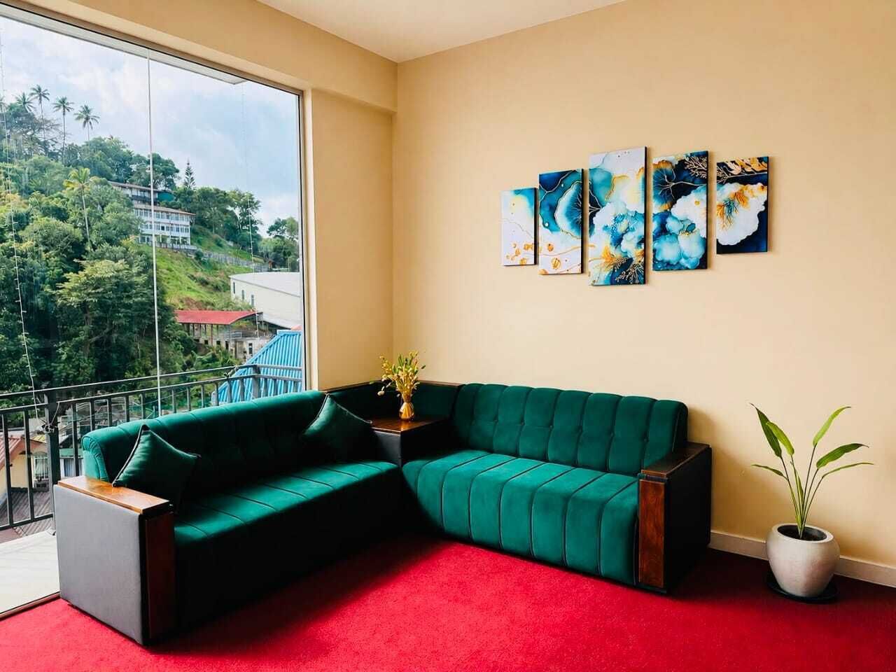A room with a green sectional sofa, a red carpet, and a large window with a view of greenery and buildings.
