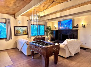 A cozy living room with a foosball table, a sofa, a fireplace, and a TV displaying a mountain scene. - Cozy place close to ski resorts, lake, town & golf (Big Bear)