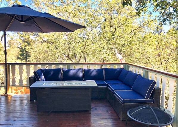 Ein überdachter Außenbereich mit einer dunklen Sofagarnitur und einem Tisch, umgeben von einer Holzterrasse und Bäumen.