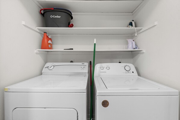 A laundry room with two white washing machines, a black bucket labeled 'O-Cedar,' and a red bottle on a shelf.