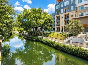 Ein Kanal mit Spiegelung von Bäumen und Gebäuden, umgeben von Grünflächen und einem modernen Wohnhaus mit Balkonen.