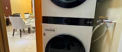A stacked washer and dryer unit with a control panel, a wall-mounted hose reel, and a tiled floor.