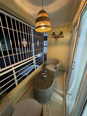 Een balkon met rieten meubilair, een hanglamp en uitzicht op gebouwen.