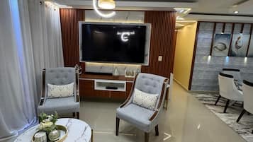 A modern living room with a flat-screen TV, two grey armchairs, a coffee table, and a dining area with white chairs.