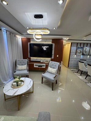Una sala de estar moderna con un televisor de pantalla plana, dos sillones grises, una mesa de centro y una zona de comedor con sillas blancas.