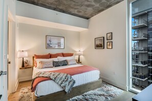 A bedroom with a bed, bedside lamps, a rug, and a window with a view of the building's exterior. - Luxury Lake View 23rd Fl Condo with Private Balcony & Rooftop Pool, Rainey St (Austin)