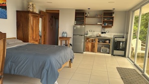 A bedroom with a bed, a kitchen with a refrigerator, stove, and cabinets, and a view through a glass door. - Studio apartment with ocean view, minutes away from the beach, surf, & swimming! (San Juan del Sur)
