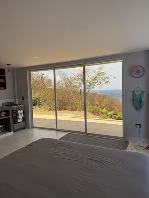 A bedroom with a large sliding glass door leading to a balcony with a view of trees and the sea. - Studio apartment with ocean view, minutes away from the beach, surf, & swimming! (San Juan del Sur)