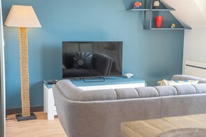 A modern living room with a flat-screen TV, a grey sofa, a tall floor lamp, and a small table with a lamp. - T2 avec aperçu mer, plage et commerces à pied (Larmor-Plage)
