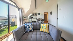 Een moderne woonkamer met een grijze bank, twee sierkussens, een groot raam en uitzicht op het omliggende landschap.