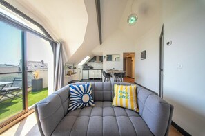 A modern living room with a grey sofa, two decorative pillows, a large window, and a view of the outside. - T2 avec aperçu mer, plage et commerces à pied (Larmor-Plage)