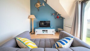 Een moderne woonkamer met een grijze bank, een flatscreen televisie op een witte dressoir, een blauwe accentmuur en een raam met gordijnen.